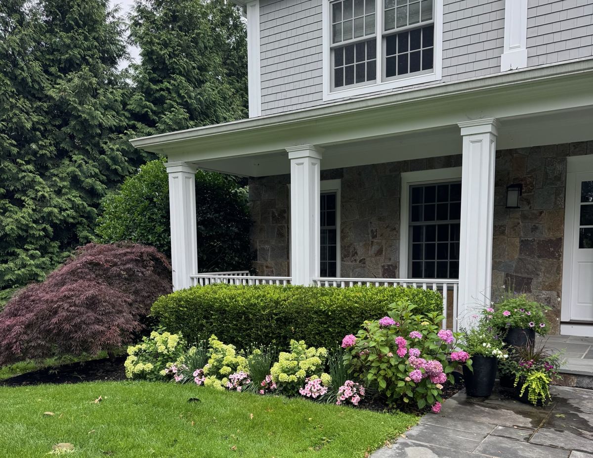 Same front porch with lush green hedge, blooming hydrangeas, and colorful potted flowers